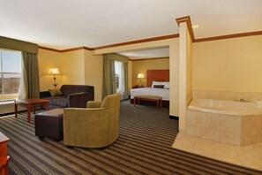 Habitación de hotel con una cama grande, un área de descanso con sofá y sillón, una mesita, un banco y un baño separado con bañera.