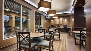 Un espace repas moderne avec des tables rondes et des chaises en bois, de grandes fenêtres équipées de stores et un téléviseur à écran plat fixé au mur.