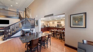 Un vaste espace de salle à manger avec une table centrale entourée de chaises, un escalier avec une rampe et, au fond, un coin cuisine.