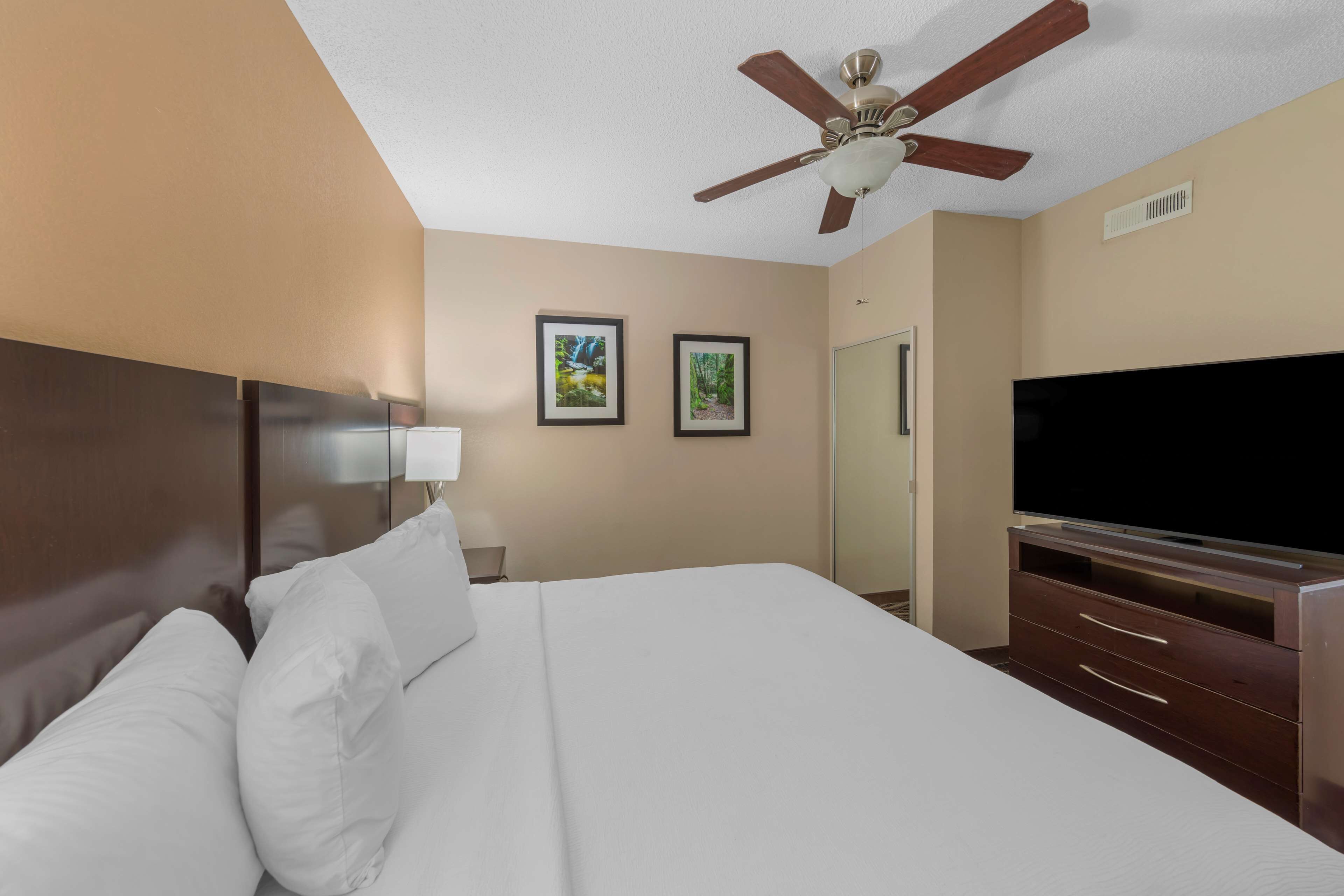Habitación de hotel con una cama grande, ventilador de techo, televisión de pantalla plana y dos cuadros en la pared.