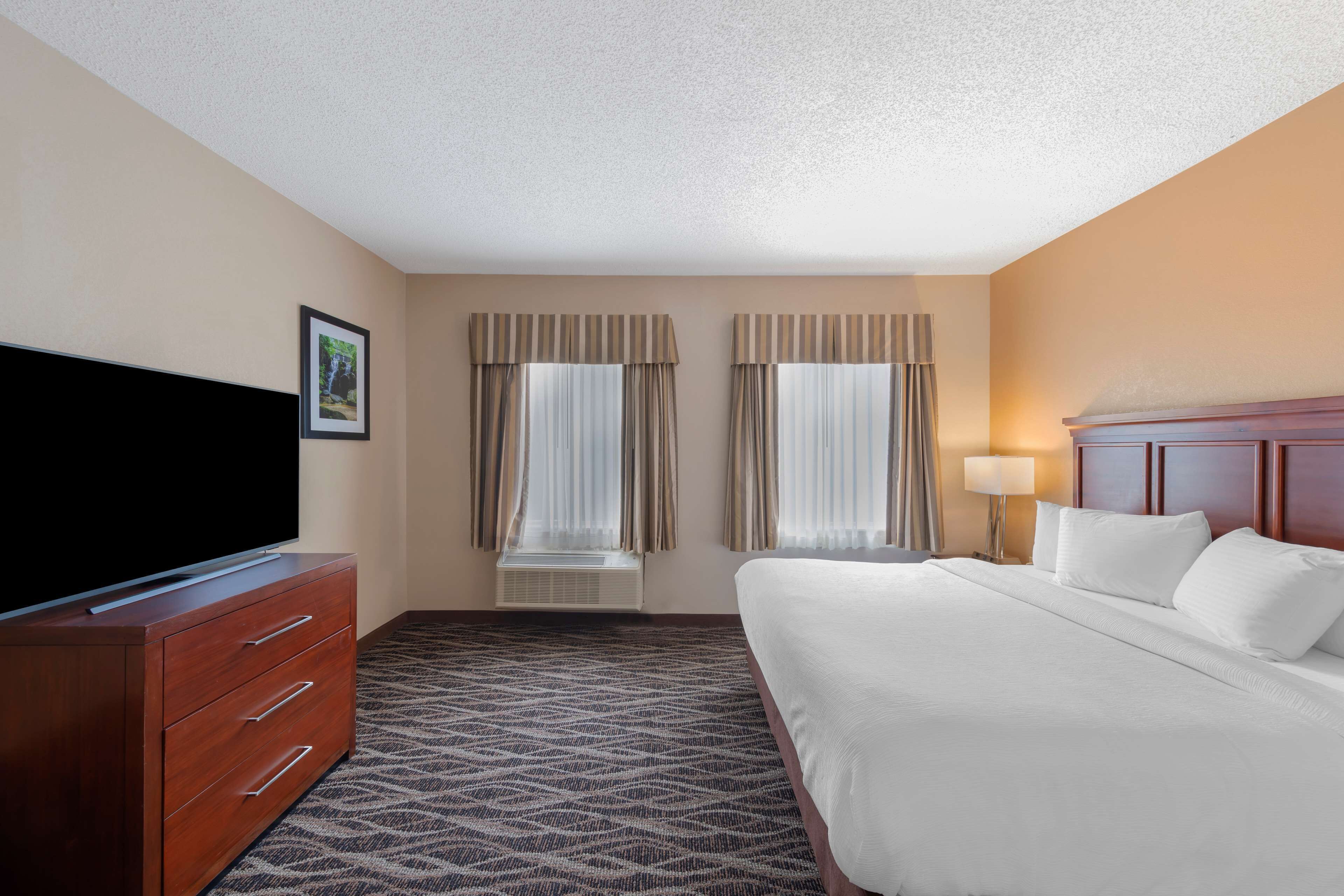 A hotel room with a bed, a TV on a wooden dresser, and two windows with curtains.