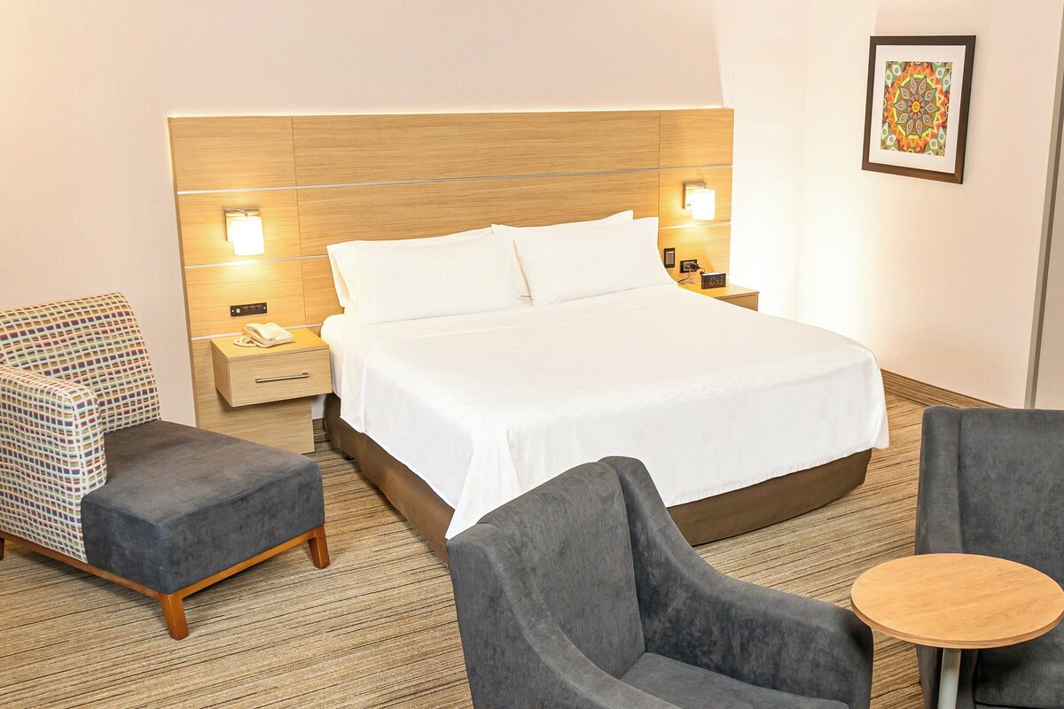 A hotel room with a large bed, two armchairs, a small round table, a nightstand with a phone, and a wall-mounted light fixture.