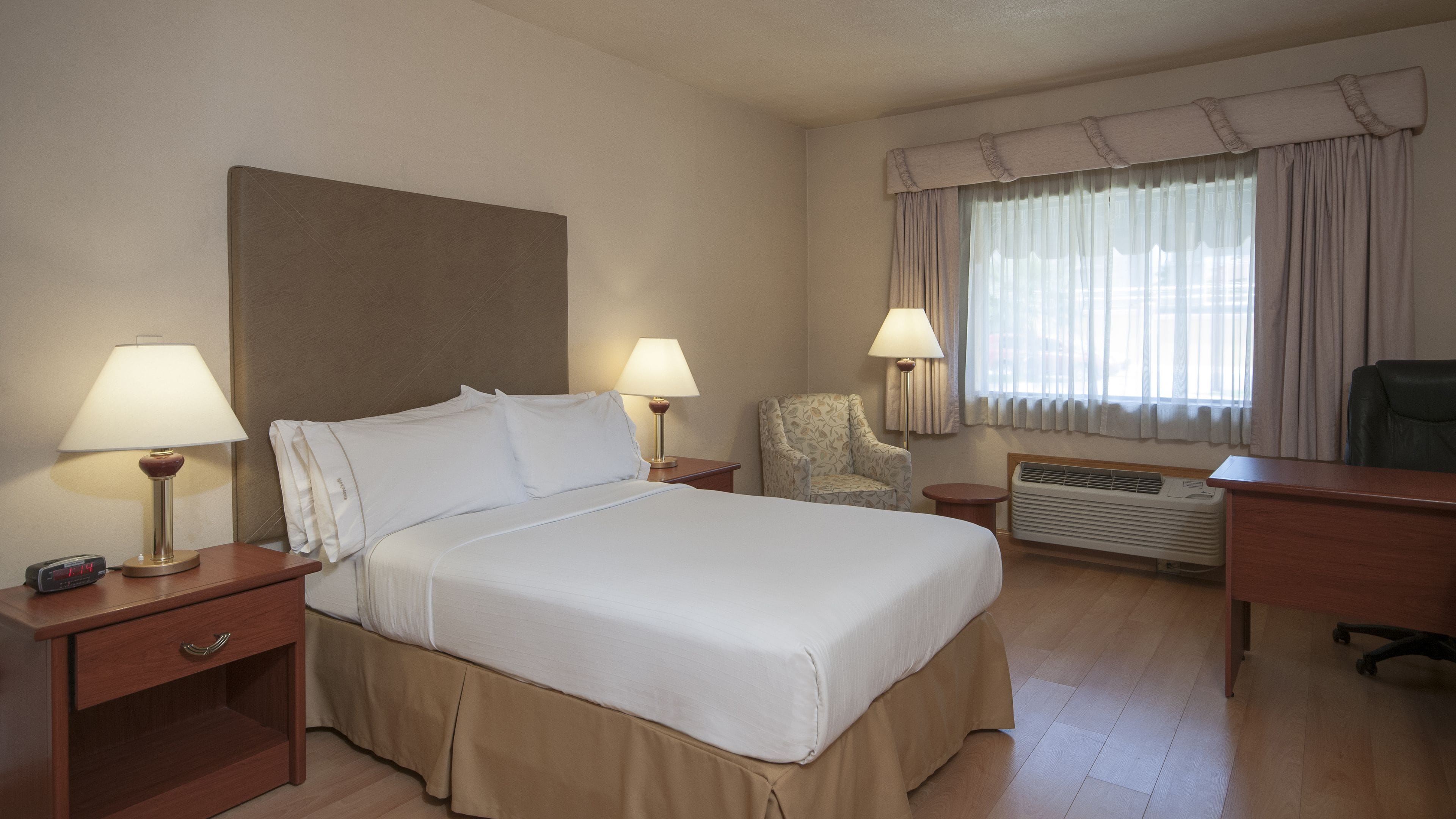 Ein Hotelzimmer mit Bett, zwei Nachttischen mit Lampen, einem Sessel, einem Schreibtisch und einem Fenster mit Vorhängen.