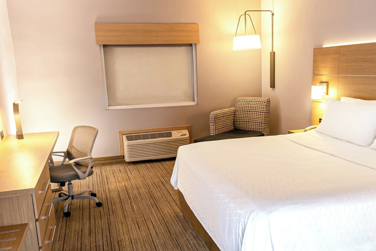 A hotel room with a bed, a desk with a chair, a wall-mounted heater, a wall-mounted lamp, and a window with blinds.