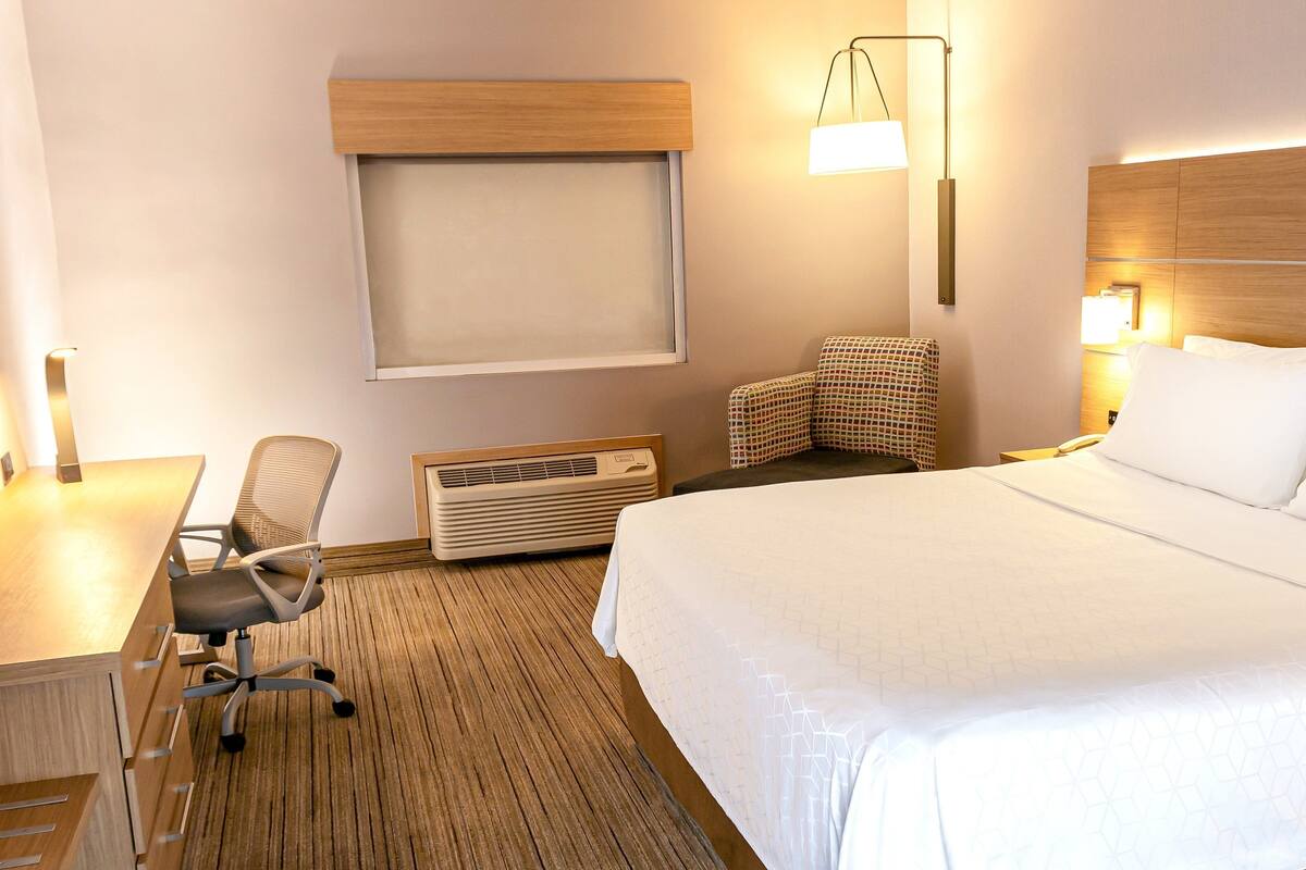 A hotel room with a bed, a desk with a chair, a wall-mounted heater, a wall-mounted lamp, and a window with blinds.