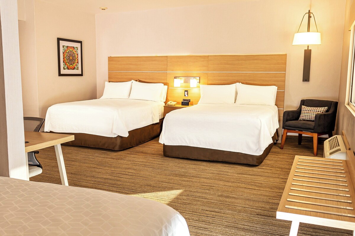 A hotel room with two beds, a wooden headboard, bedside lamps, a chair, and a small table.
