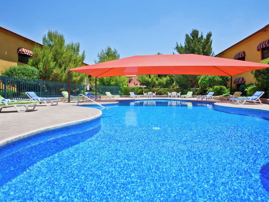 A swimming pool with multiple lounge chairs and umbrellas, surrounded by greenery and buildings.