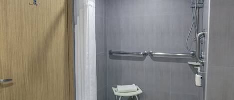 Zona de ducha con barra de apoyo, un taburete pequeño con una toalla y un cabezal de ducha.
