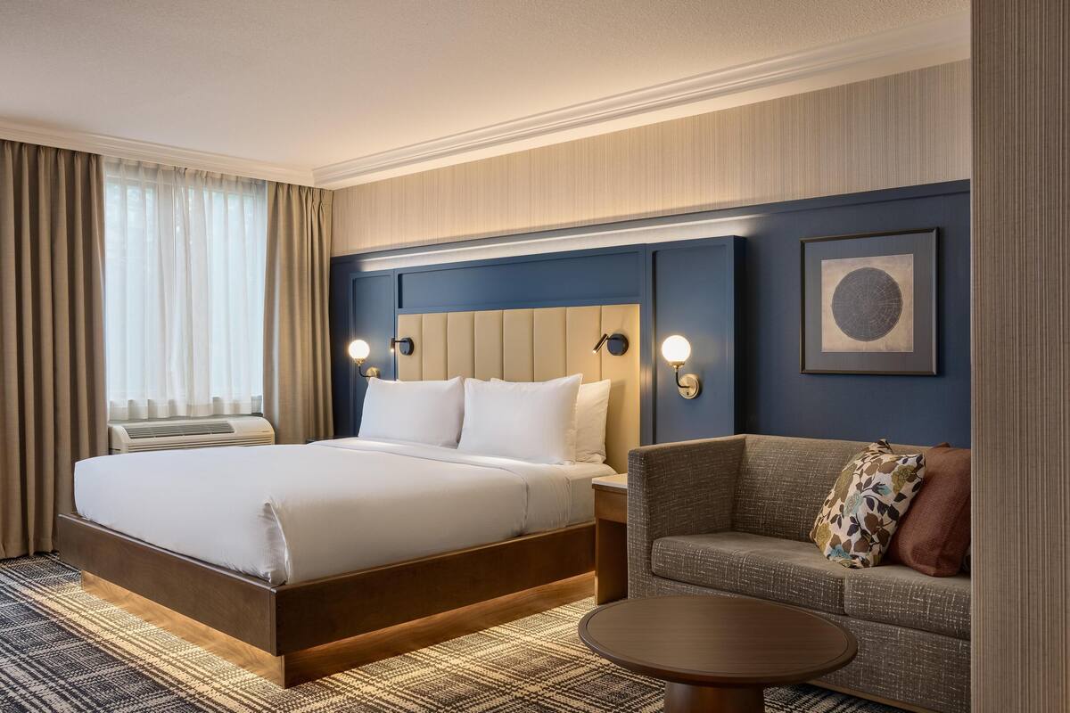 A hotel room with a large bed, a sofa, a small table, and a wall-mounted artwork.