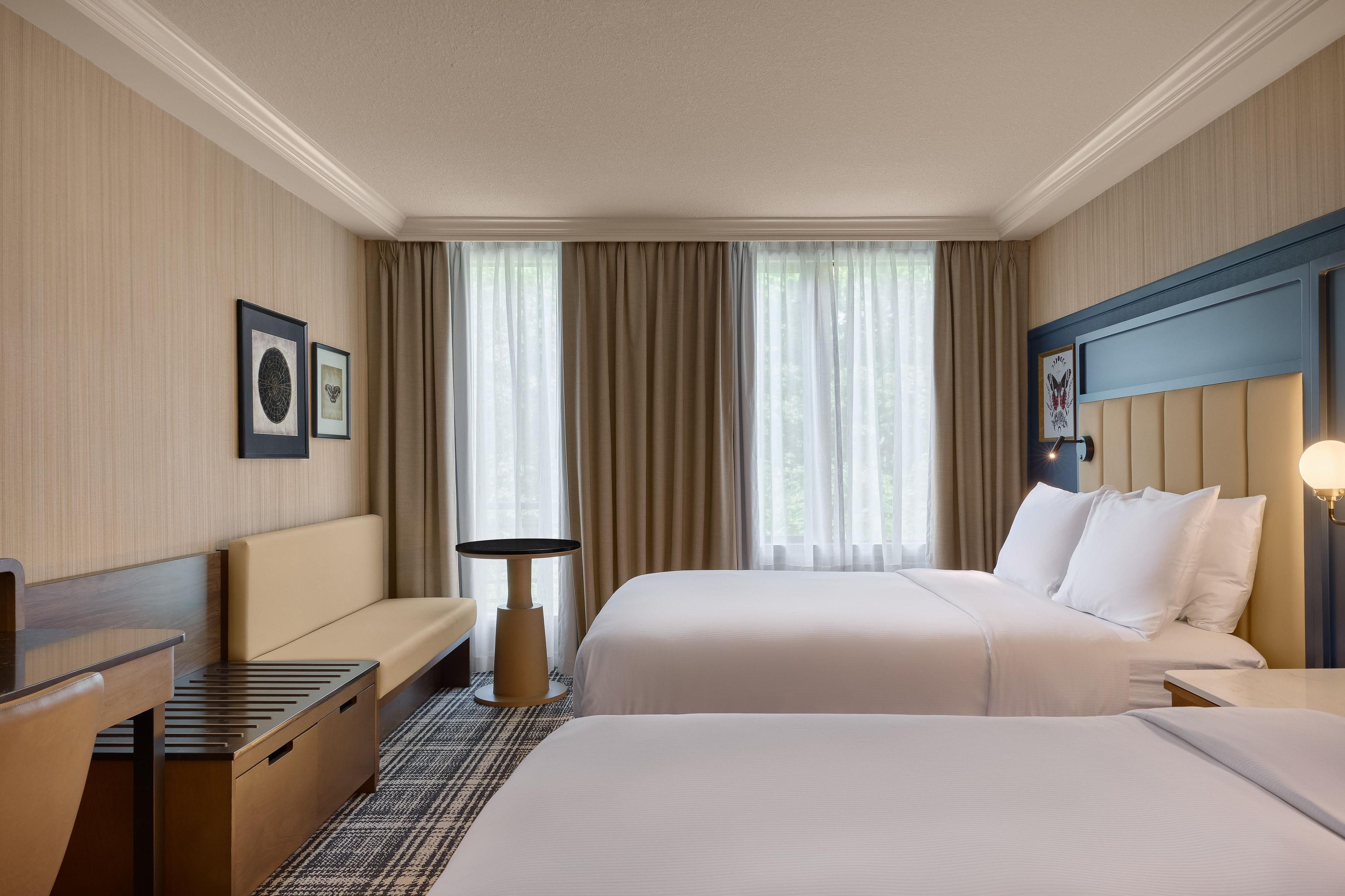 A hotel room with two beds, a desk, a chair, a small table, and a window with curtains.