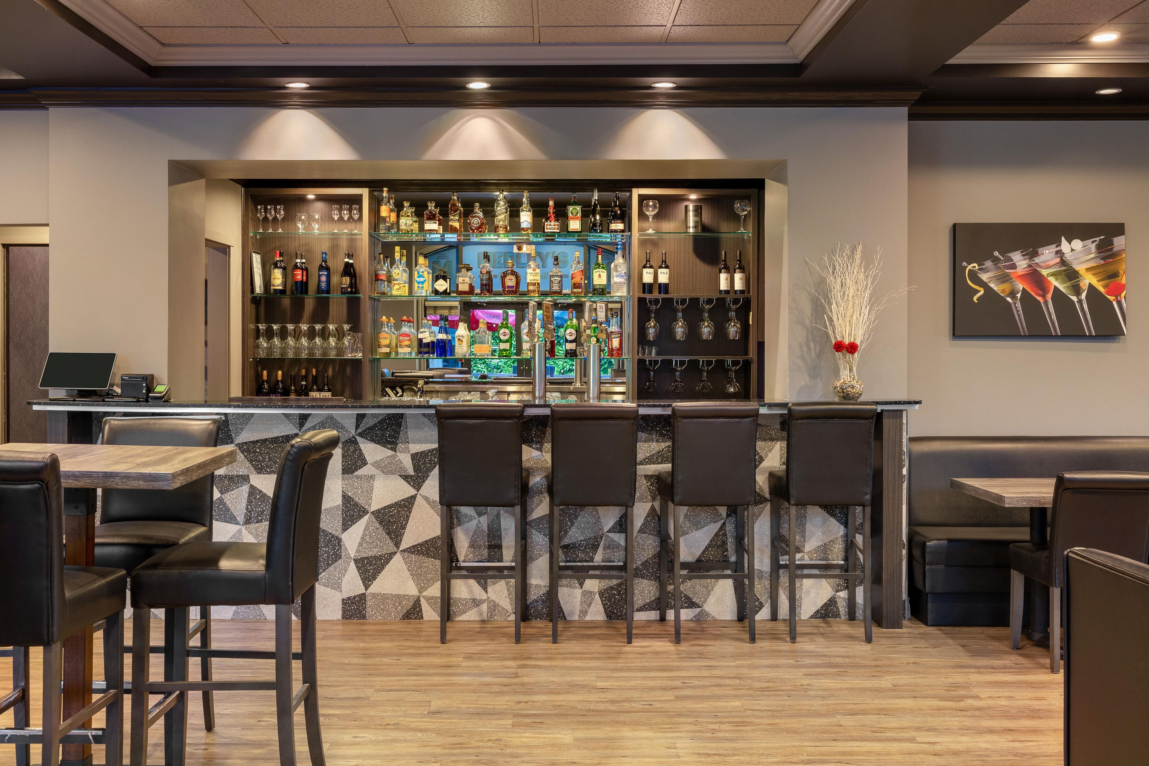 A bar area with a counter, shelves of bottles, glasses, and a seating area with chairs and tables.