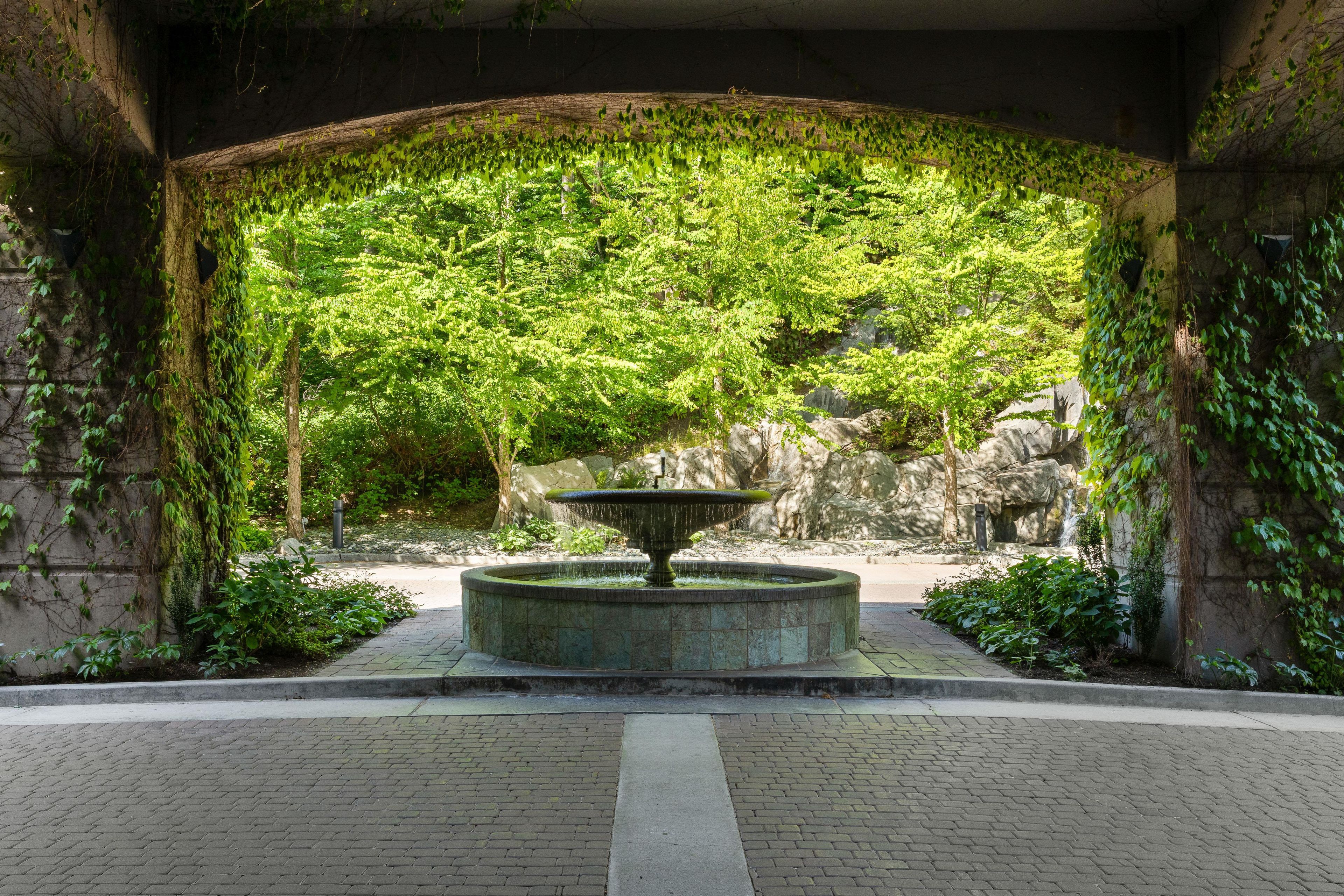 A stone fountain in a covered garden area with lush greenery and a stone wall.