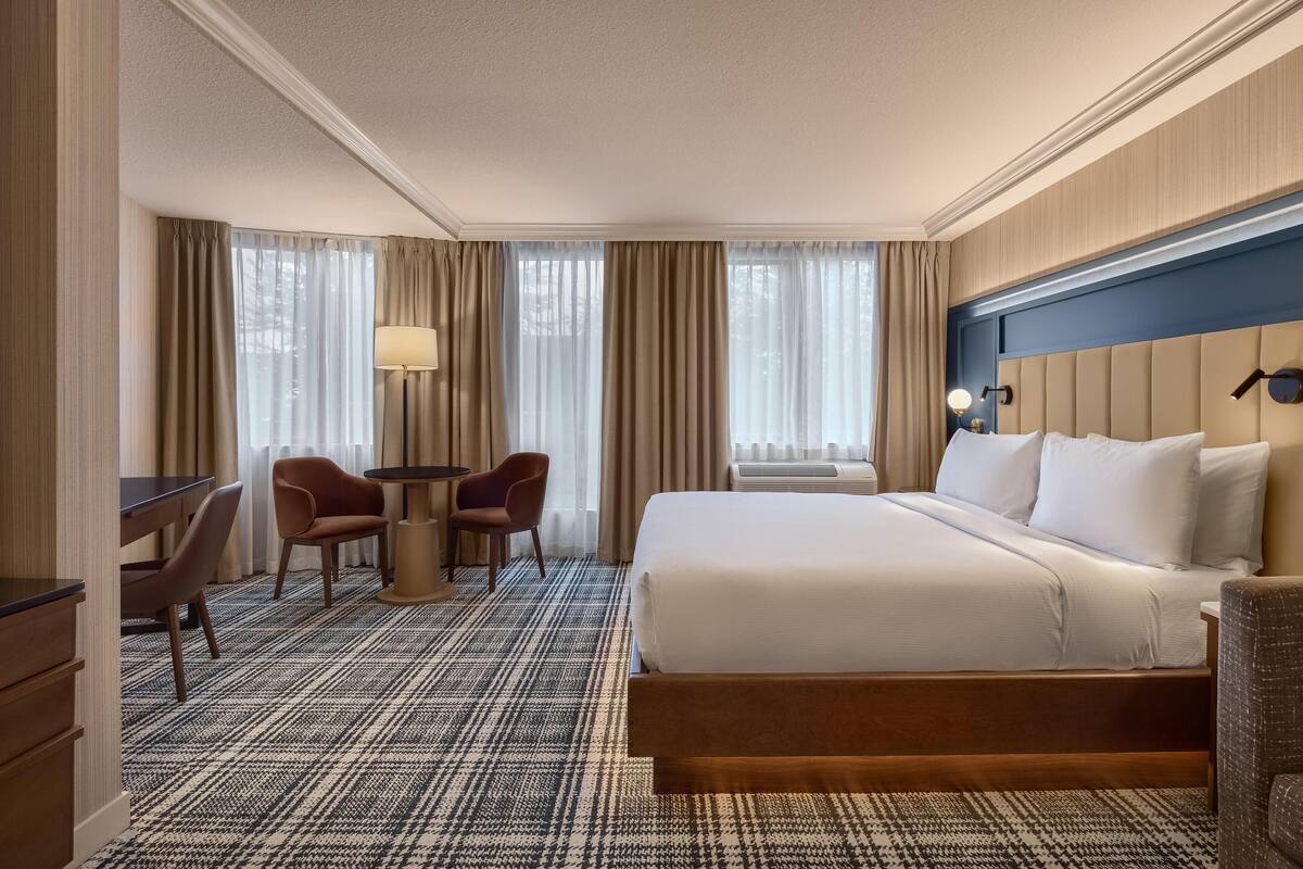 A hotel room with a large bed, a desk, two chairs, a lamp, and a view through the window.