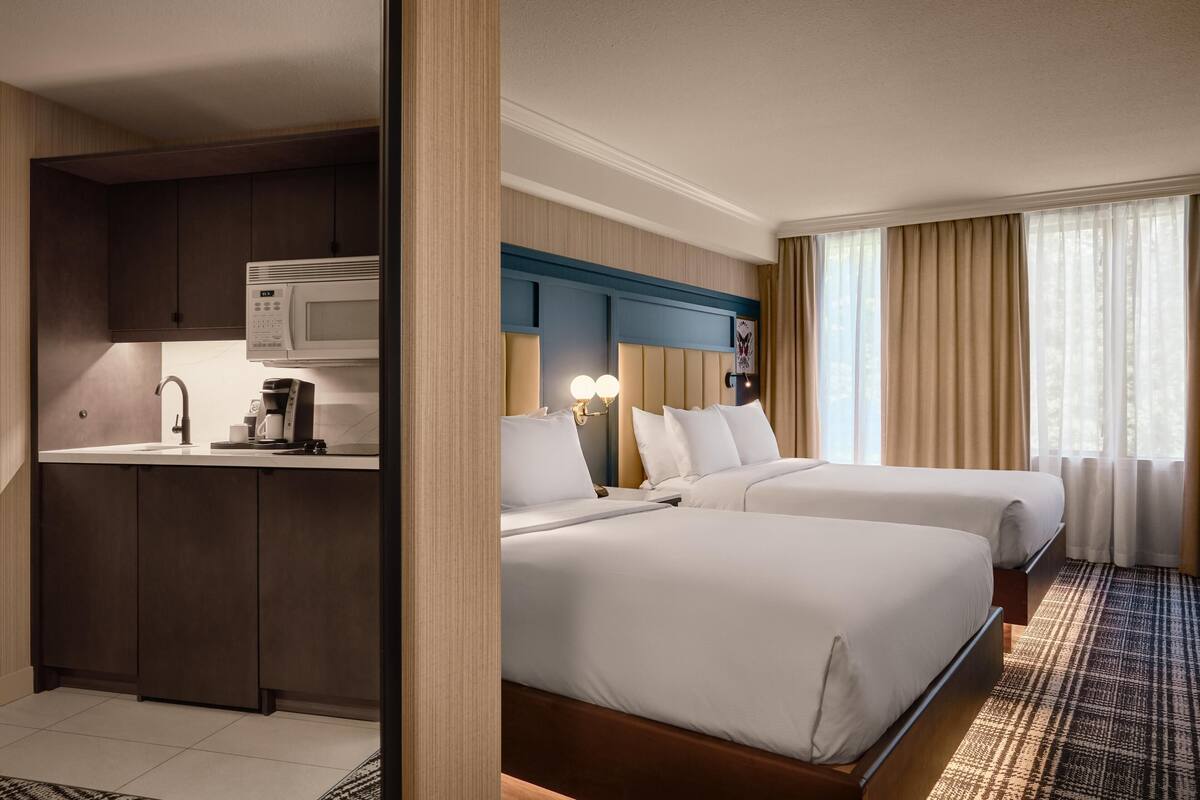A hotel room with a kitchenette, two beds, a large window with curtains, and a patterned carpet.