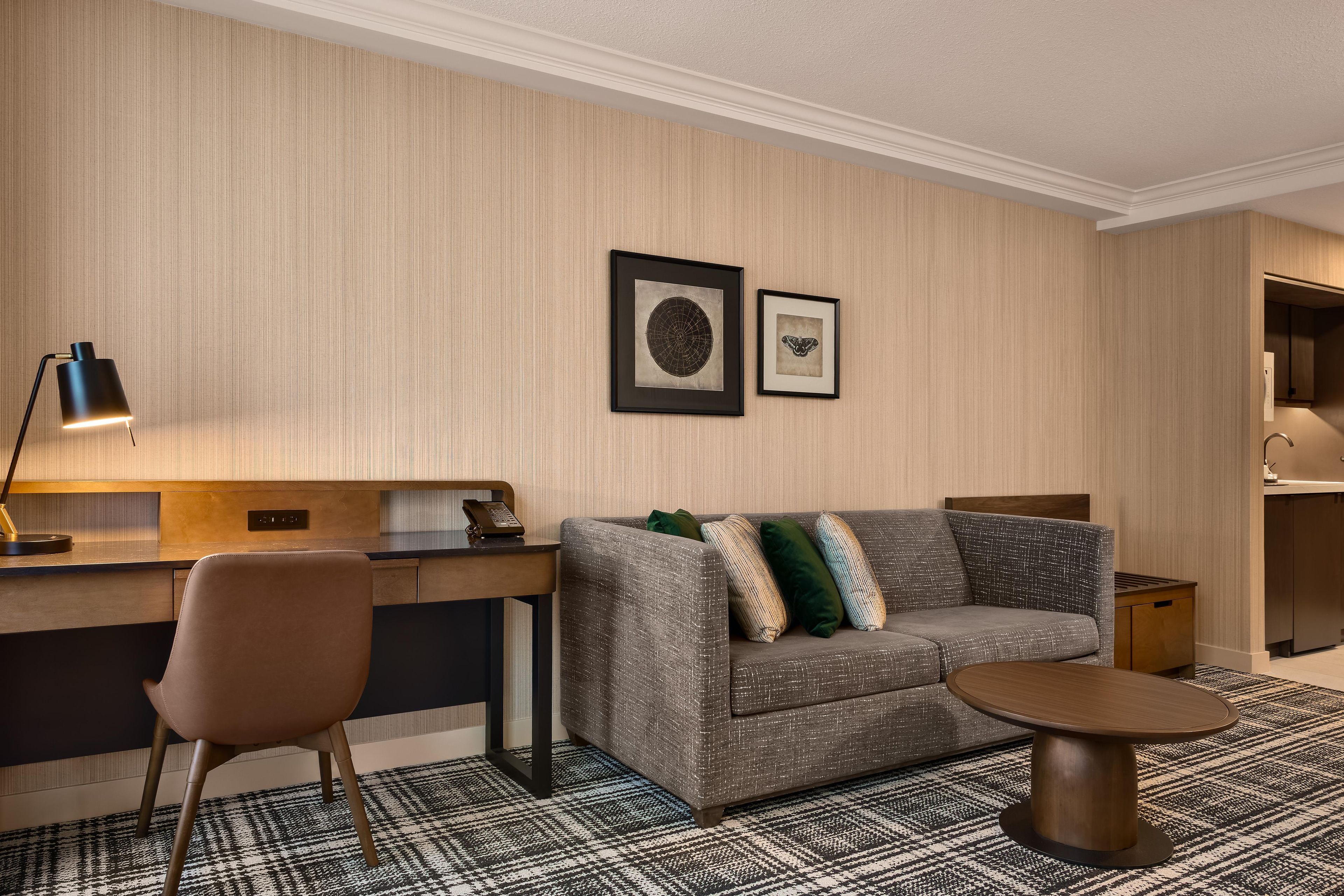 Et hotelværelse med sofa, et lille bord, et skrivebord, en stol, en lampe og billeder i rammer på væggen.
