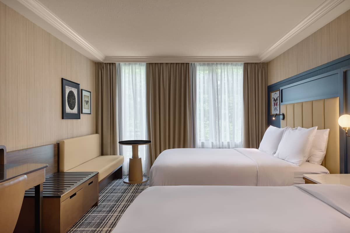 A hotel room with two beds, a desk, a chair, a small table, and a window with curtains.