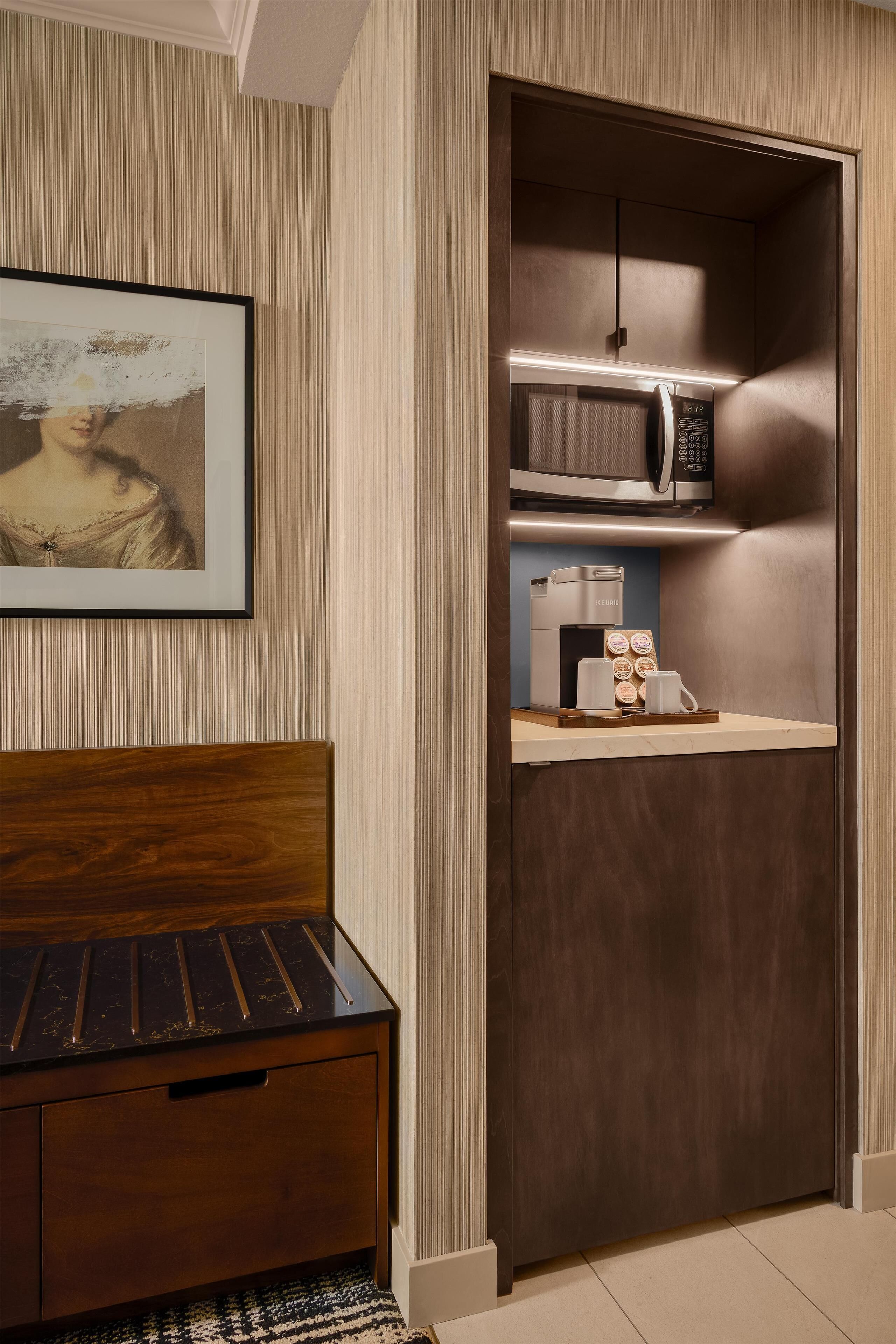 A compact room with a bed, a small wooden cabinet, a microwave, a coffee maker, and a framed picture on the wall.