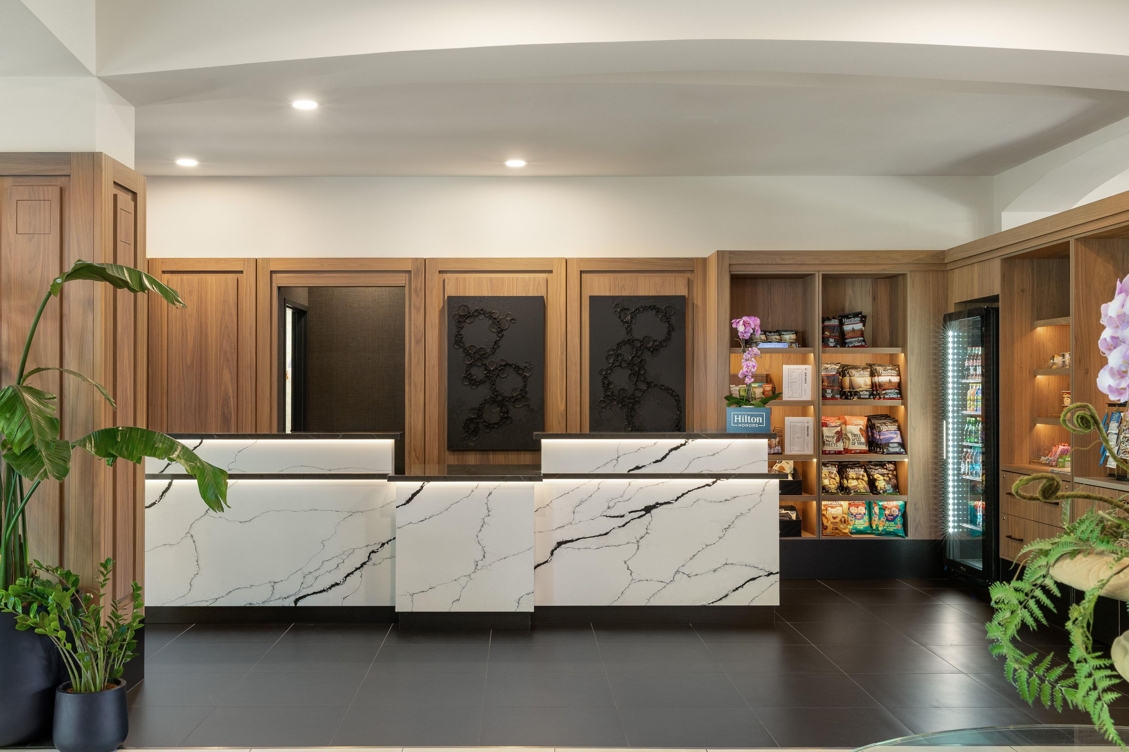 Un moderno vestíbulo de hotel con mostradores de recepción de mármol, muebles de madera y una vitrina refrigerada.