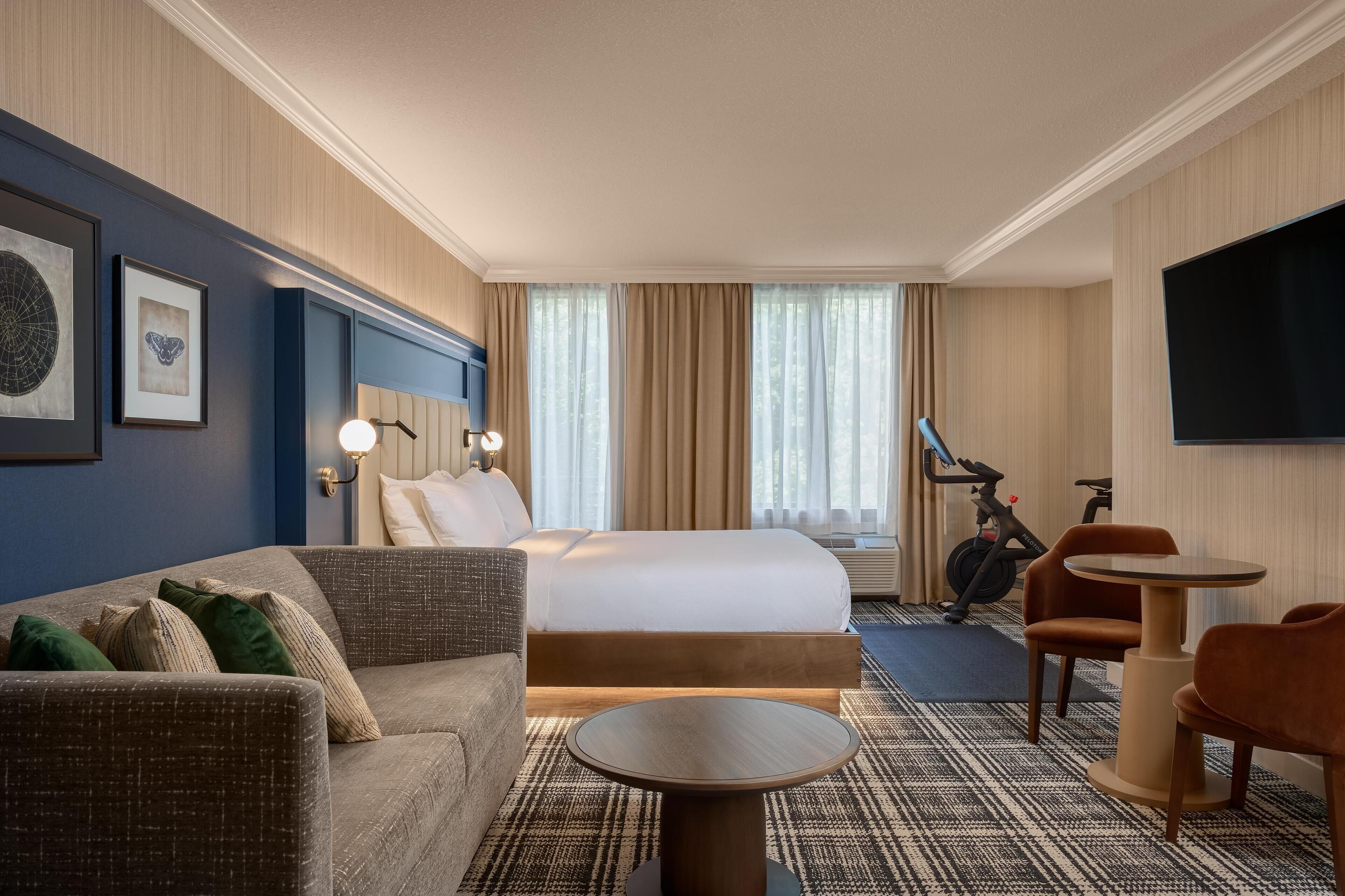 A hotel room with a bed, a sofa, a small round table, a TV, and exercise equipment.