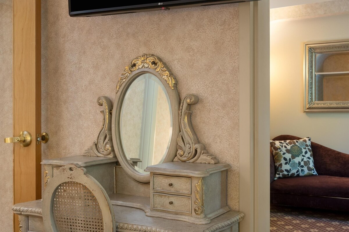 A room with a vanity, a television mounted above it, a mirror, a door, and a patterned carpet.