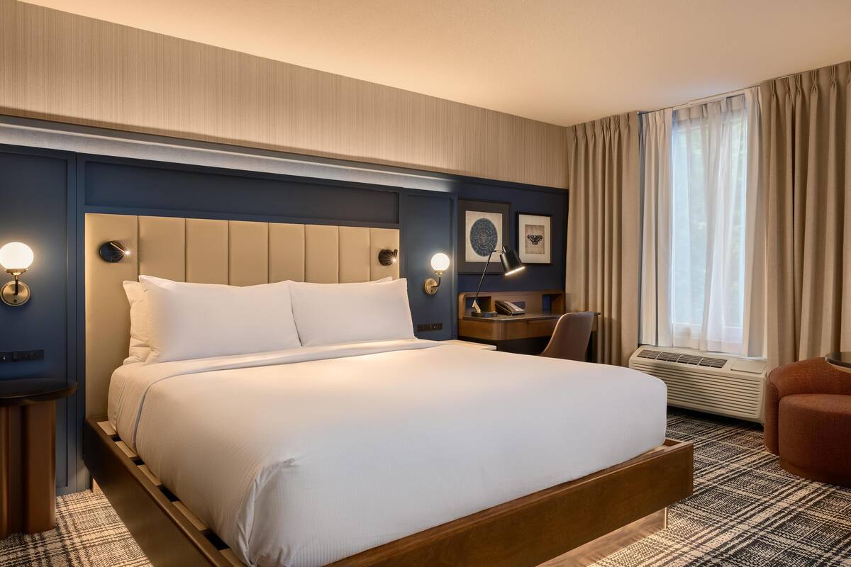 A hotel room with a large bed, bedside tables, a desk, a chair, and a window with curtains.