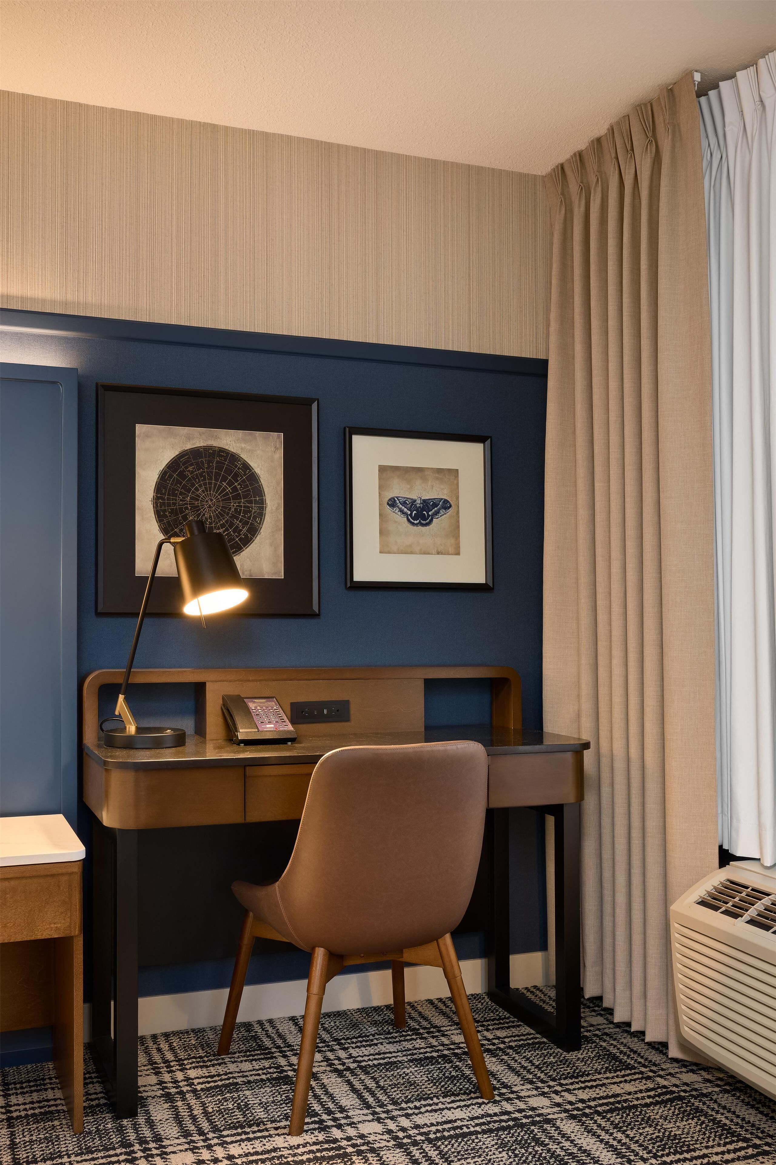 A hotel room with a desk, chair, framed artwork, and a window with curtains.