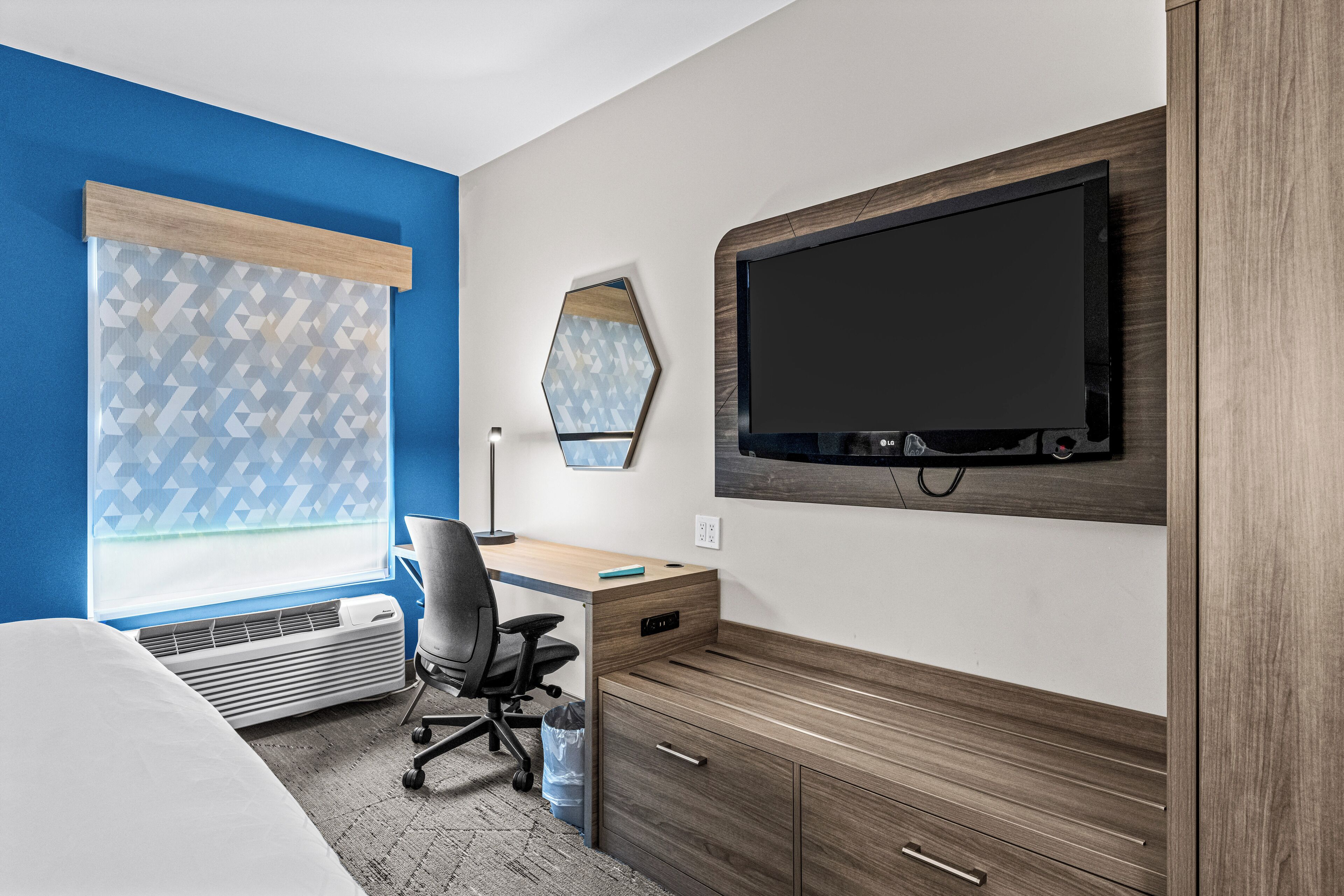 A hotel room with a bed, a desk, a chair, a TV mounted on the wall, and a window with patterned curtains.