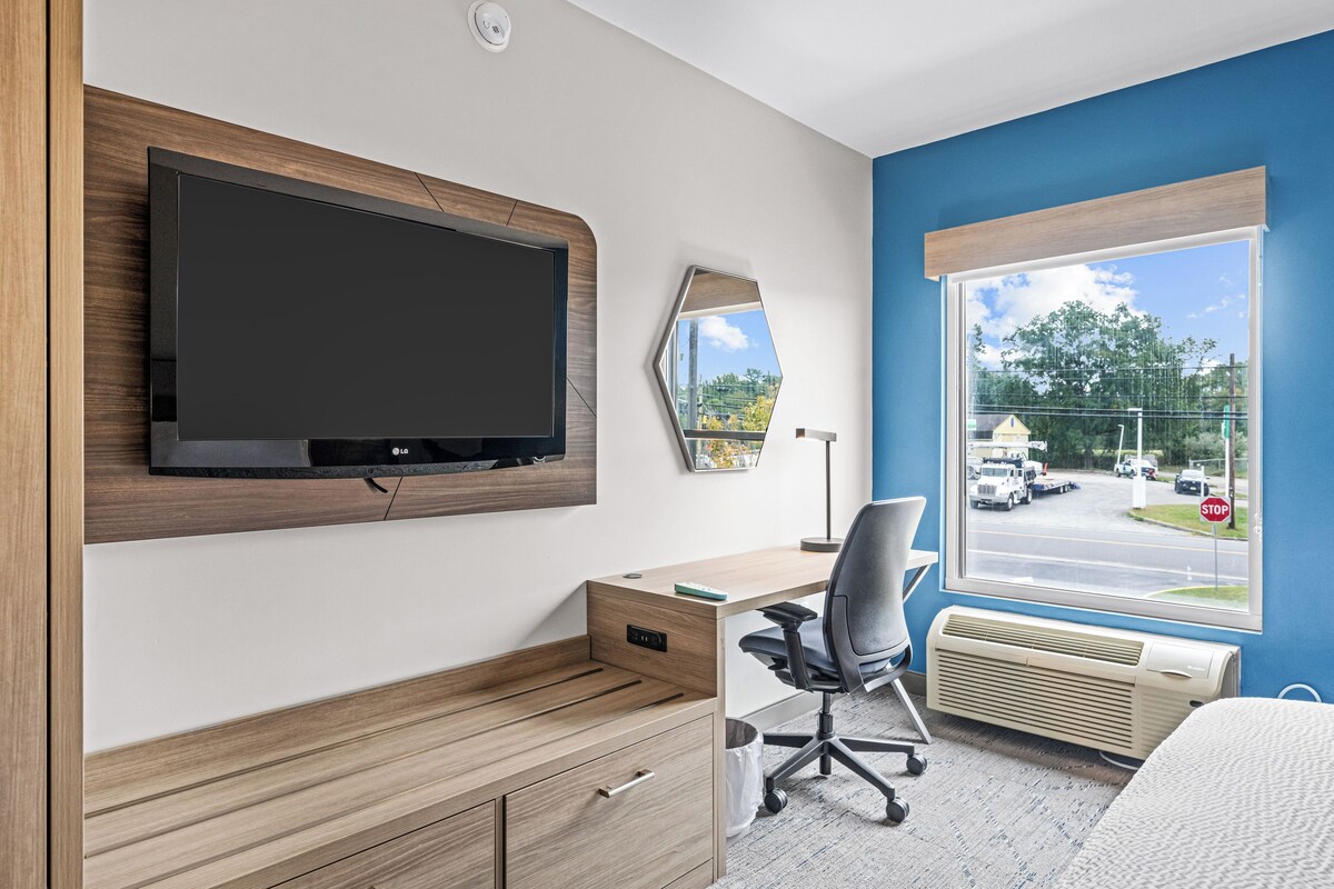 Een moderne hotelkamer met een flatscreen televisie aan de wand, een houten bureau met stoel, een groot raam met uitzicht op de straat en een radiator onder het raam.