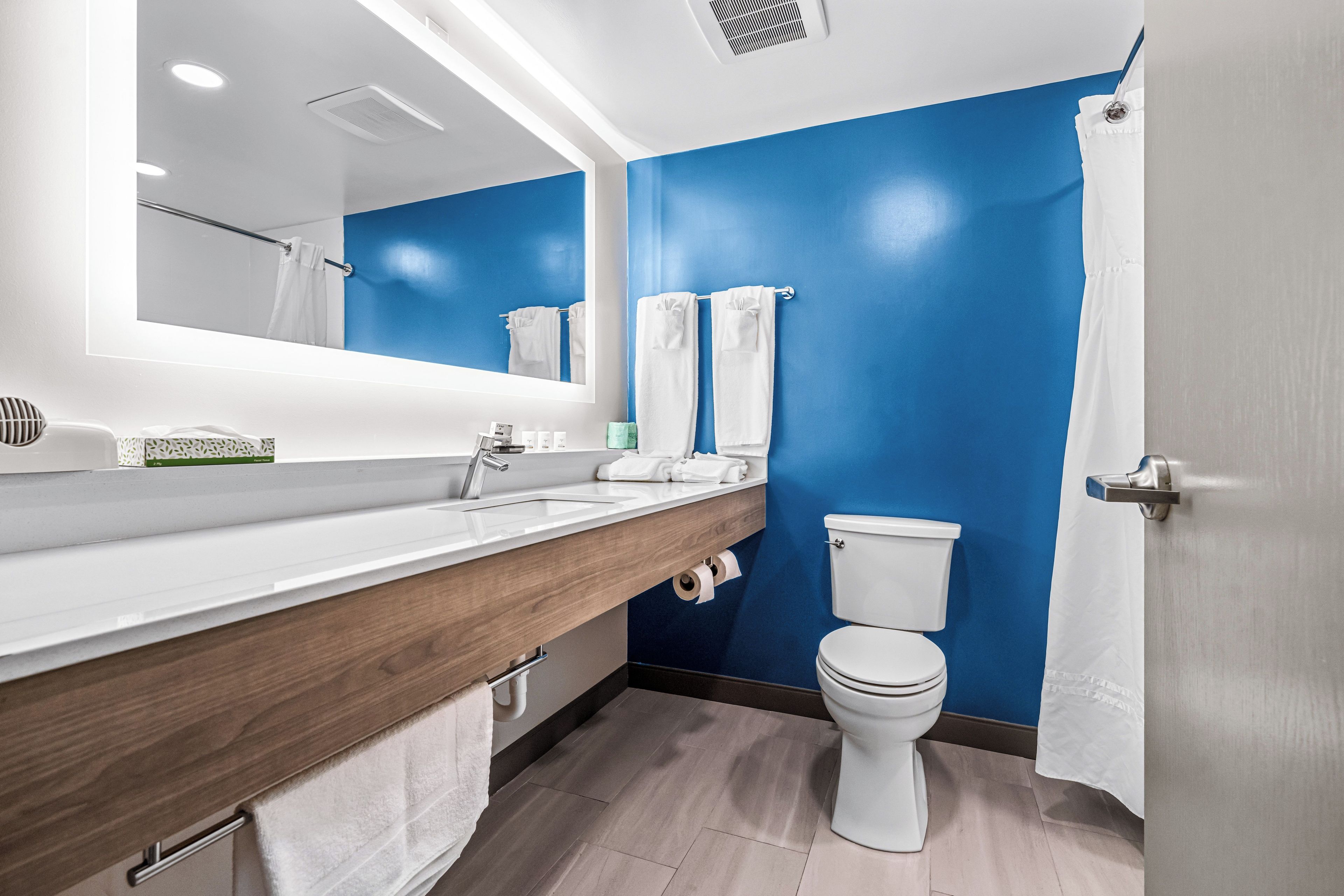 A modern bathroom with a blue accent wall, a white toilet, a wooden vanity, and a large mirror.