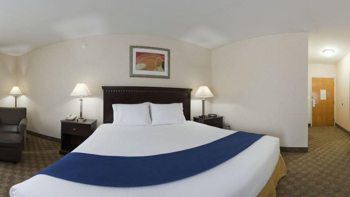 Habitación de hotel con una cama grande, dos mesitas de noche con lámparas, una silla y una puerta al fondo.
