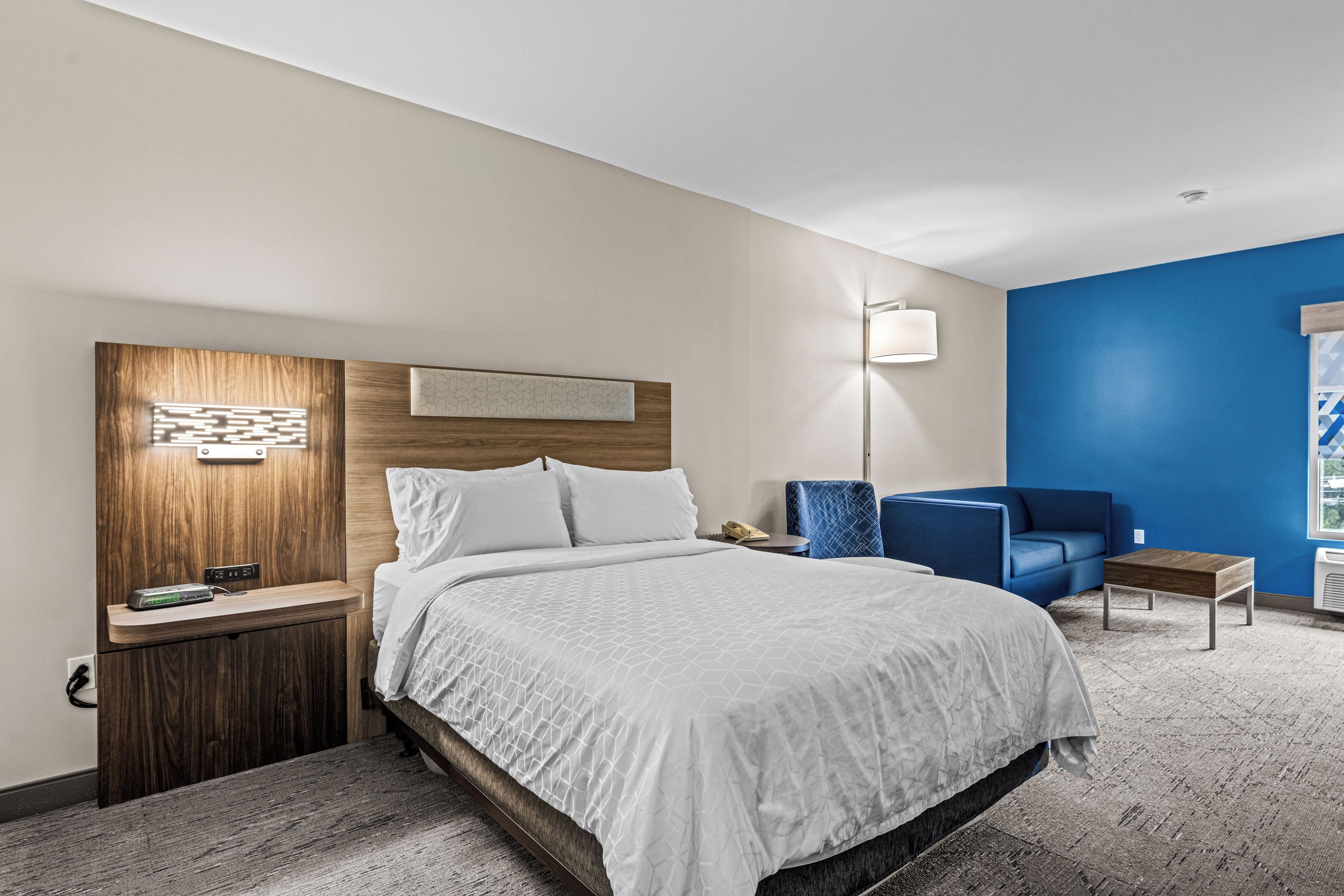 A hotel room with a large bed, a wooden headboard, a blue armchair, a small table, and a nightstand with a lamp.