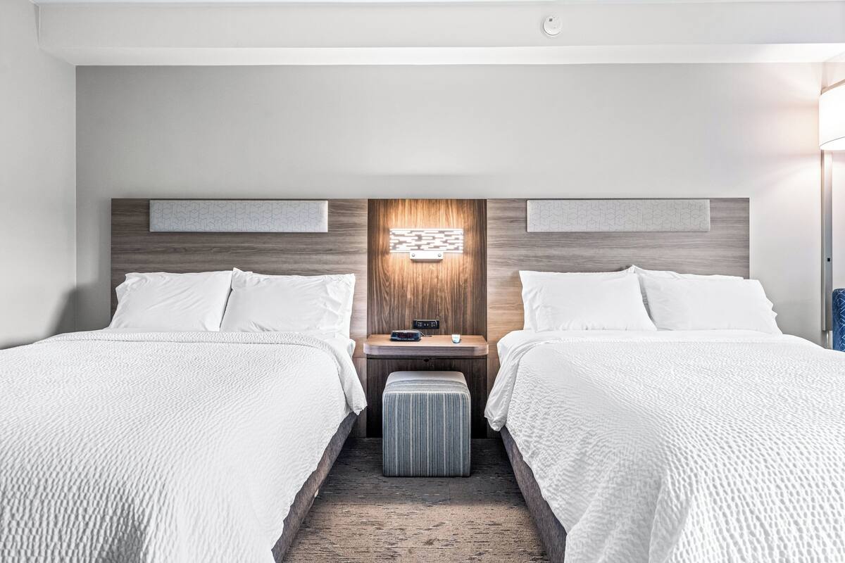 A hotel room with two beds, a nightstand with a lamp, and a wall-mounted light fixture.