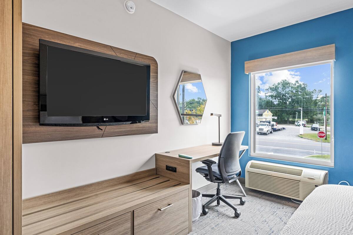 A modern hotel room with a flat-screen TV mounted on the wall, a wooden desk with a chair, a large window with a view of the street, and a radiator under the window.