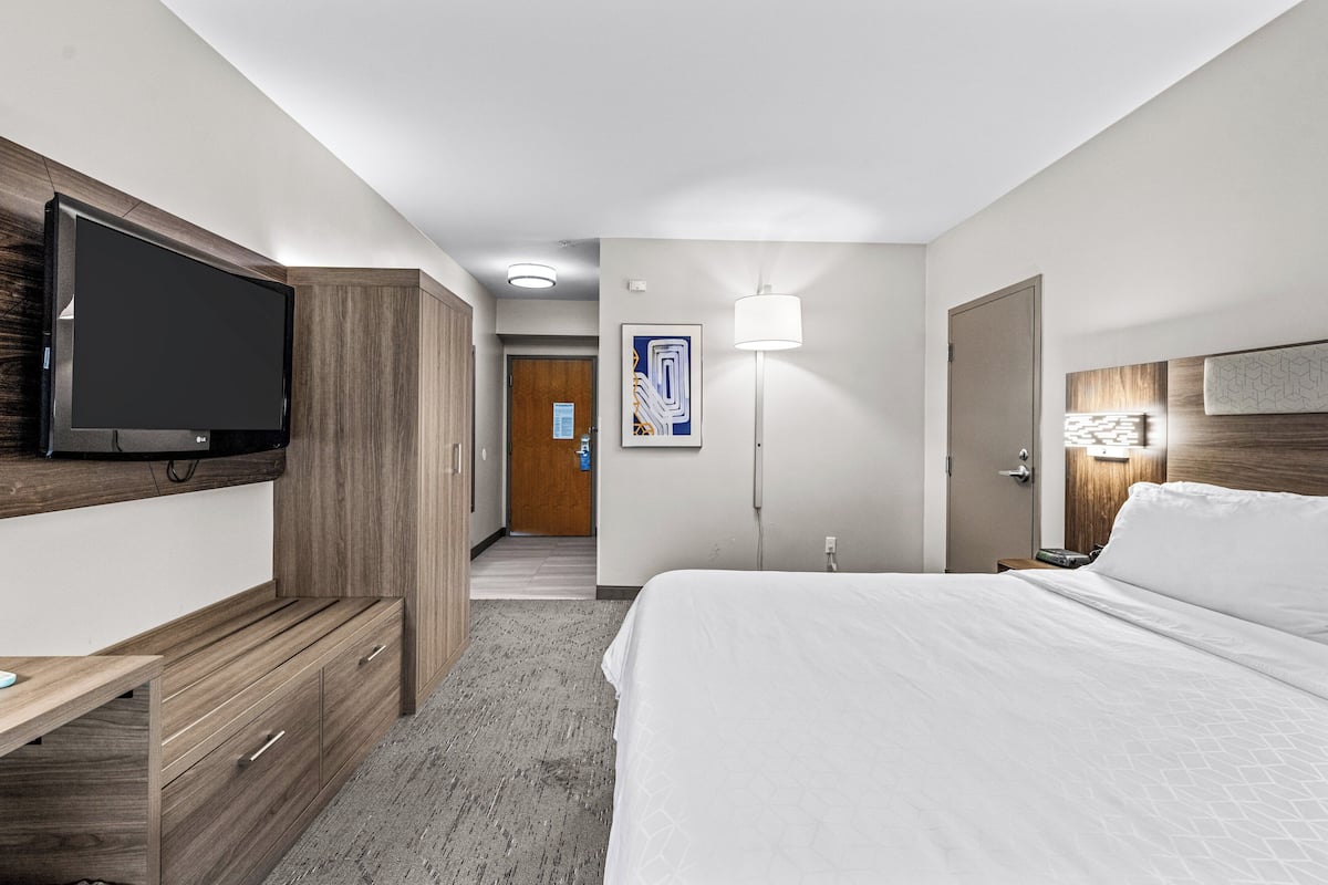 A hotel room with a bed, a desk, a television mounted on the wall, a door, and a wall-mounted light fixture.