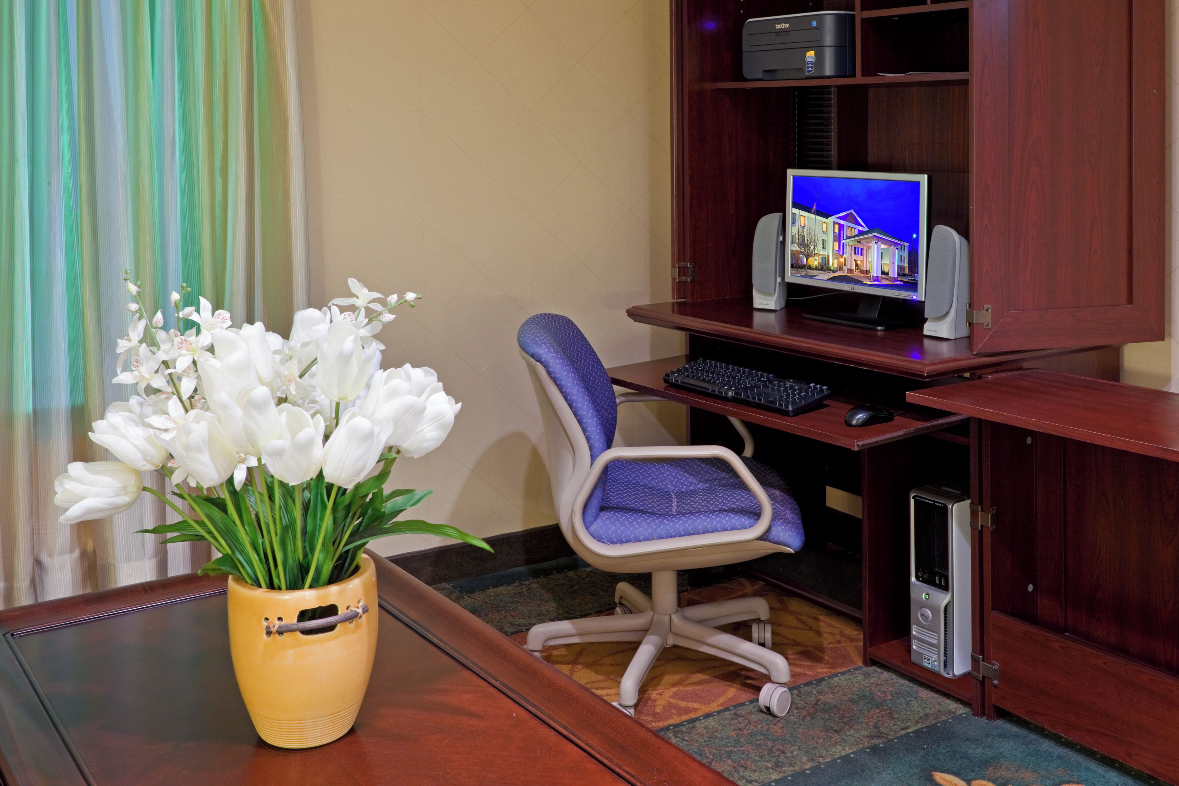 A wooden desk with a computer, a keyboard, and a mouse. There is a vase with white flowers on the desk.