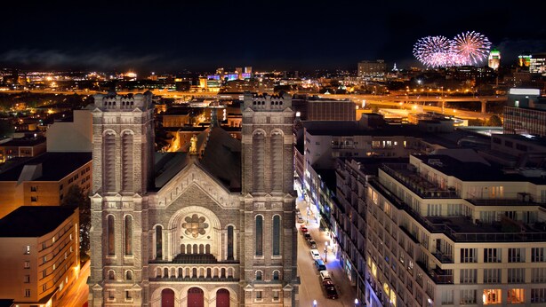 A historic church with a central dome and two towers, surrounded by modern buildings and a busy street at night.