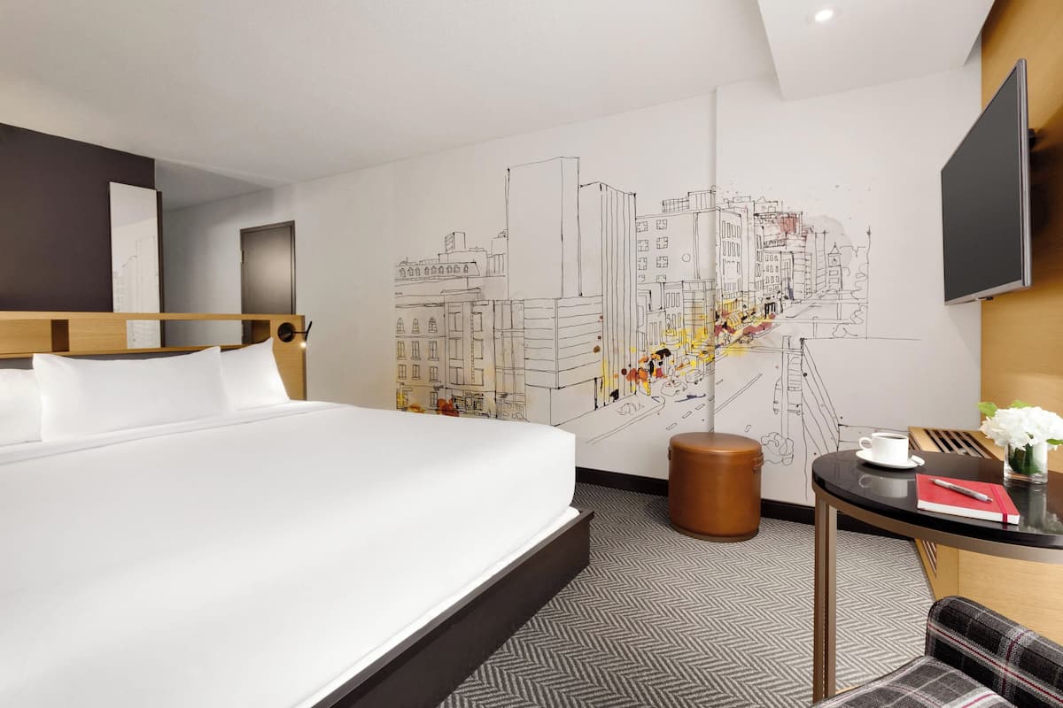 Une chambre d’hôtel avec un grand lit, une fresque représentant un paysage urbain sur le mur, une petite table ronde avec une tasse et un livre, et un téléviseur fixé au mur.