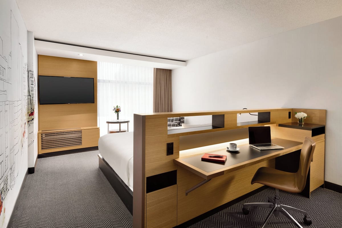 Une chambre d’hôtel avec un bureau en bois, un ordinateur portable, une chaise, un lit, une télévision et une petite table sur laquelle est posé un vase de fleurs.