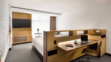 A hotel room with a wooden desk, a laptop, a chair, a bed, a TV, and a small table with a vase of flowers.