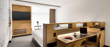 Habitación de hotel con un escritorio de madera, una computadora portátil, una silla, una cama, un televisor y una mesita con un jarrón de flores.