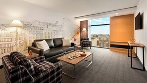 A modern hotel room with a black leather sofa, plaid armchairs, a wooden coffee table, and a cityscape mural on the wall.