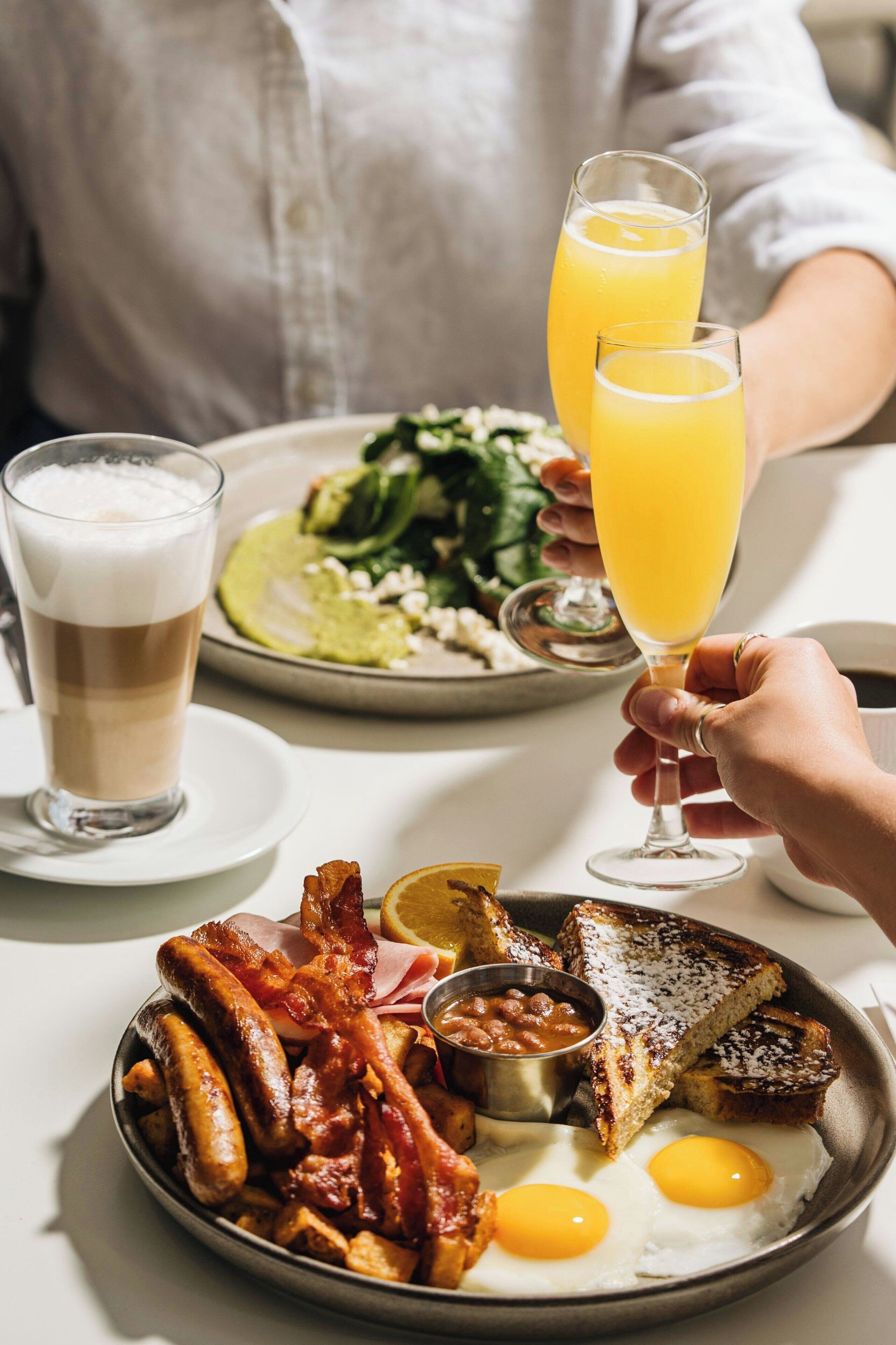 Un desayuno con un plato de huevos, tocino, salchichas, tostadas y frijoles, acompañado de dos vasos de jugo de naranja y una taza de café.
