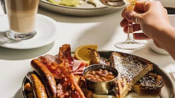 Un desayuno con un plato de huevos, tocino, salchichas, tostadas y frijoles, acompañado de dos vasos de jugo de naranja y una taza de café.