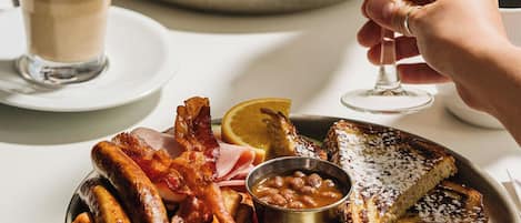 A breakfast setting with a plate of eggs, bacon, sausages, toast, and beans, accompanied by two glasses of orange juice and a cup of coffee.