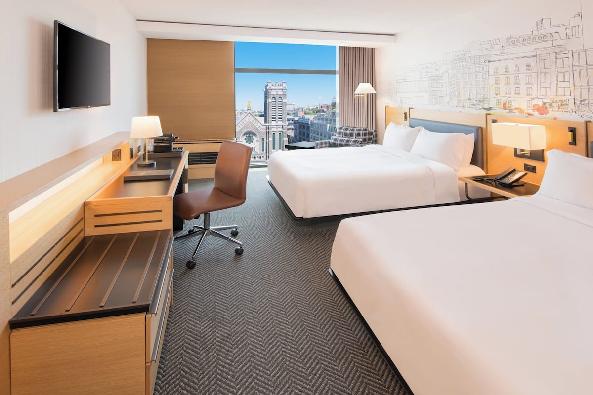 Une chambre d’hôtel avec deux lits, un bureau avec une chaise, un téléviseur à écran plat et une grande fenêtre donnant sur la ville.
