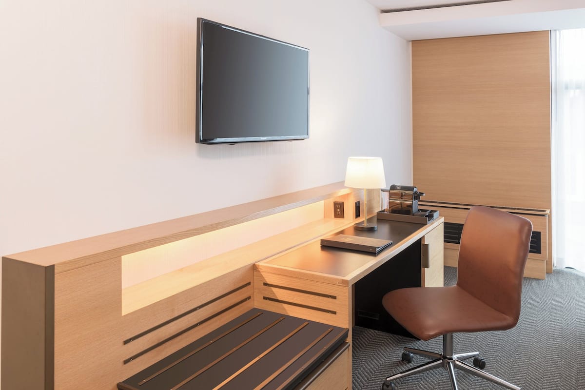 Une chambre d’hôtel moderne avec un bureau en bois, un téléviseur à écran plat fixé au mur, une chaise et une lampe.