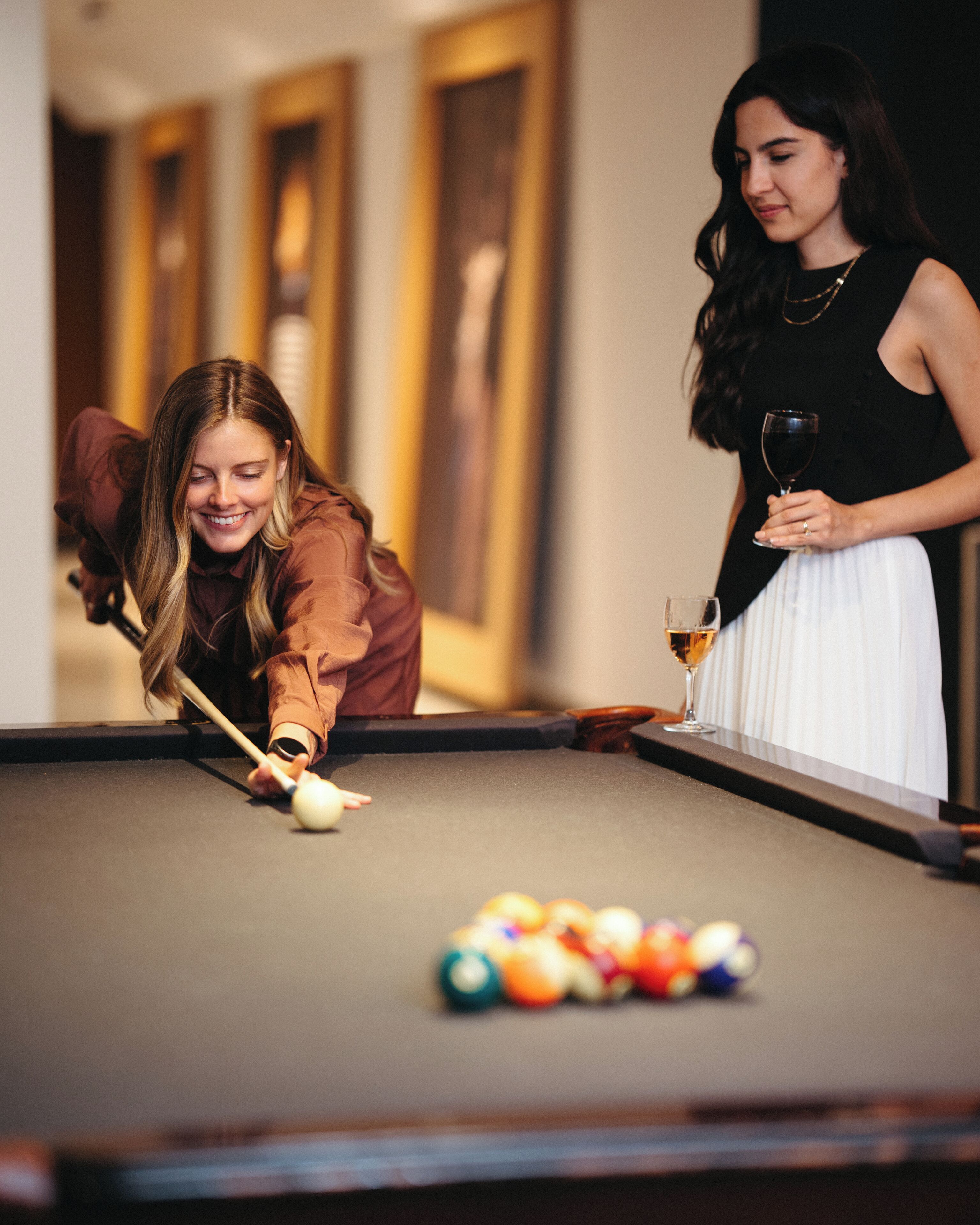 Two people playing pool in a room with a pool table and framed artwork on the wall.