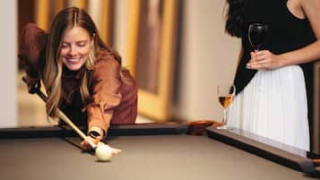 Two people playing pool in a room with a pool table and framed artwork on the wall.