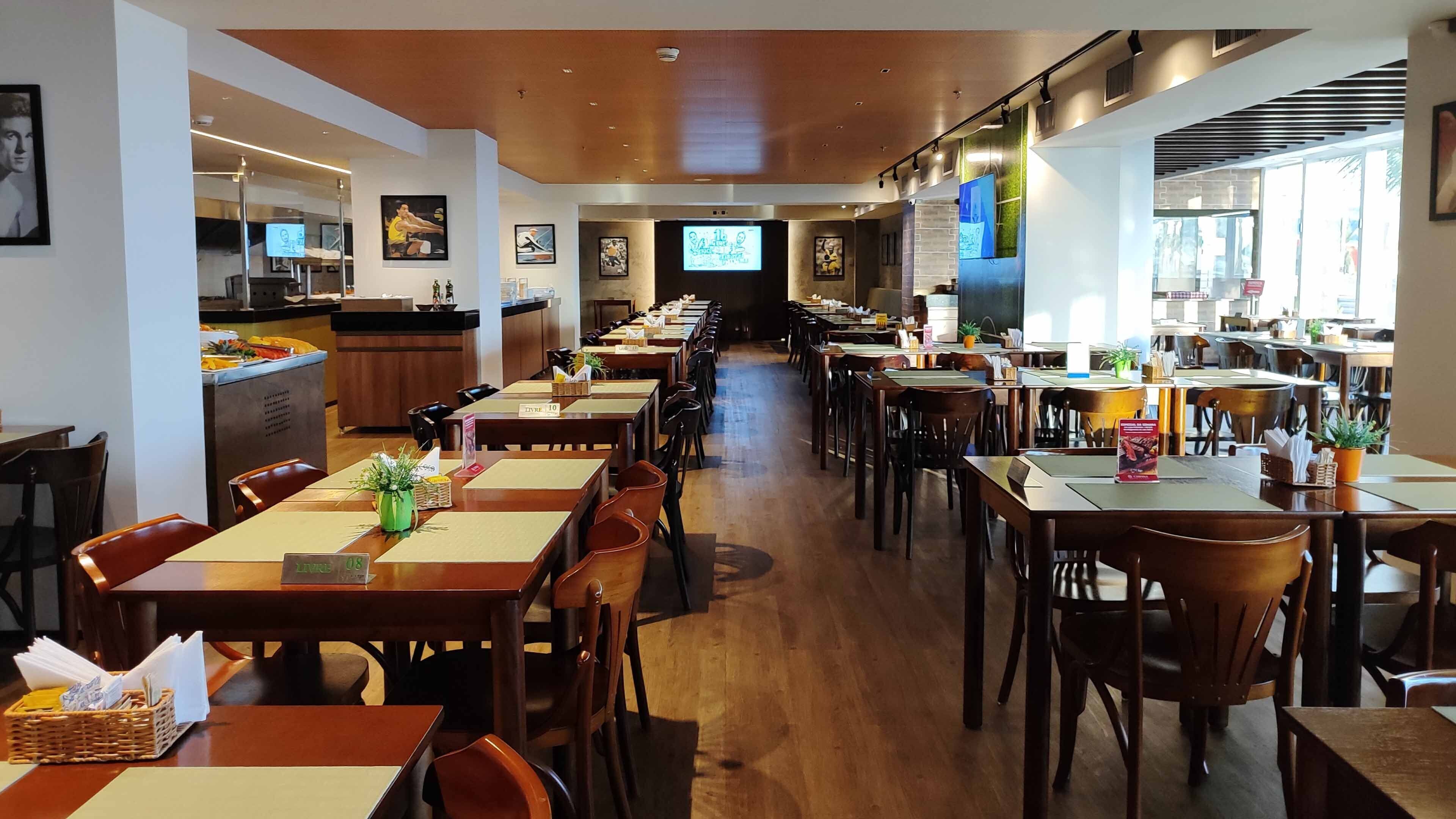 A spacious restaurant with multiple dining tables and chairs, a kitchen area, and a television screen in the center.
