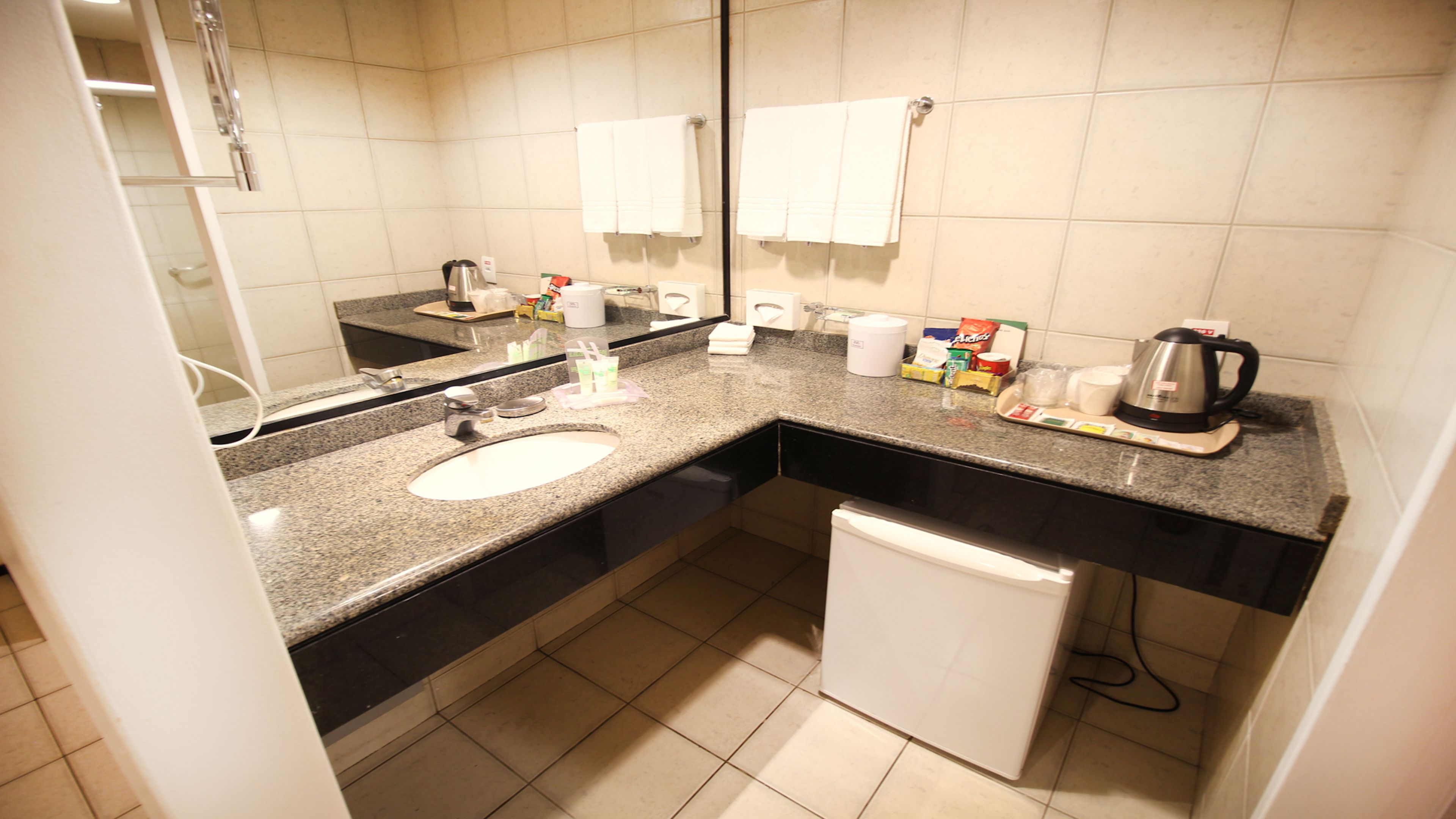 A compact hotel bathroom with a sink, mirror, towel rack, and a coffee maker.